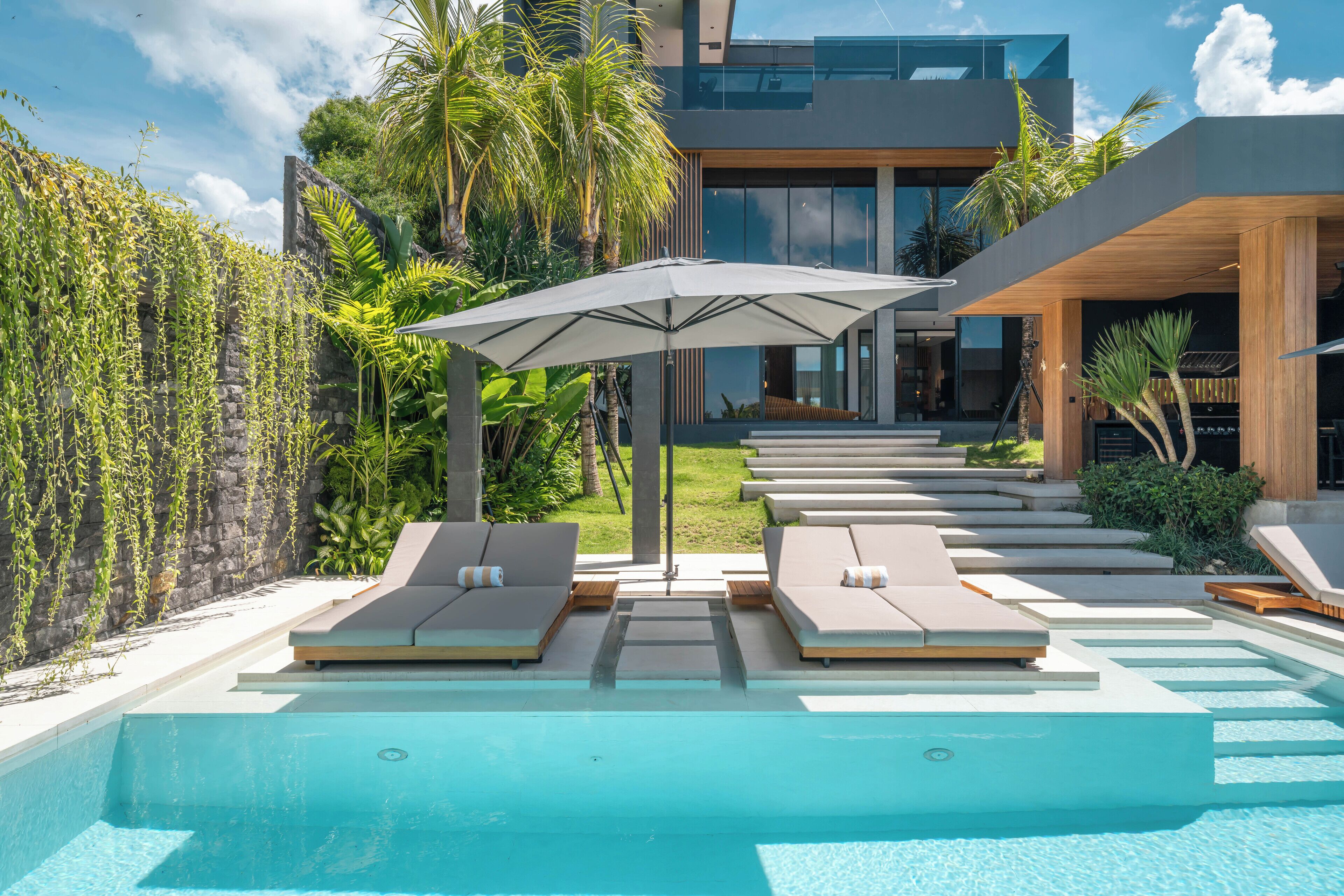 Un área de piscina moderna con sillones de descanso y una sombrilla grande, rodeada de exuberante vegetación y un edificio contemporáneo al fondo.