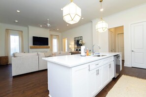 Un vaste espace de vie comprenant un grand canapé blanc, un îlot de cuisine avec un évier et des luminaires suspendus.