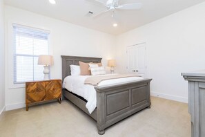 Une chambre à coucher comprenant un lit, des tables de chevet, une commode, un ventilateur de plafond et une fenêtre avec des stores.