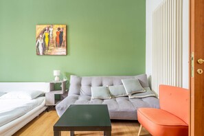 A room with a grey sofa, an orange chair, a small table, a painting on the wall, and a bedside table with a lamp.