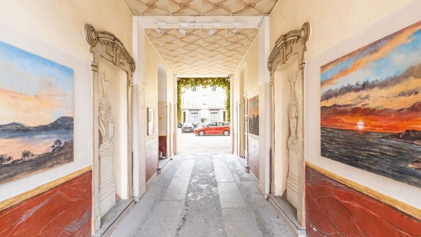 Un couloir avec un plafond carrelé, des œuvres d’art encadrées accrochées aux murs et une vue sur un bâtiment à travers une arche.