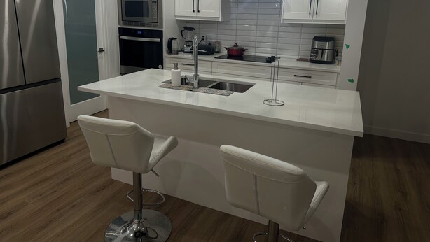 A modern kitchen with a central island, stainless steel appliances, white cabinetry, and two bar stools. - Spacious 3-Bedroom Apartment in  – Perfect for Groups & Families! (Calgary)