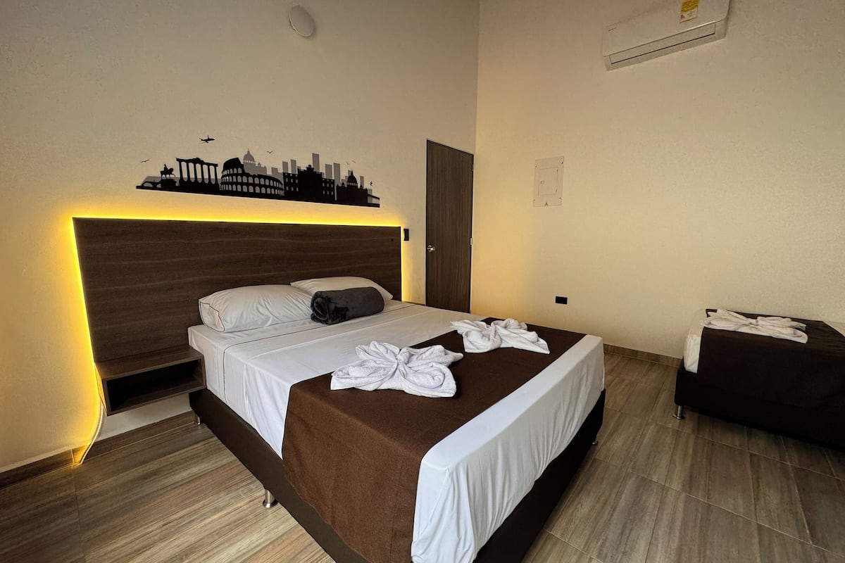 A modern hotel room with a large bed, a headboard with built-in lighting, a cityscape artwork above the bed, and a small seating area with a brown sofa.
