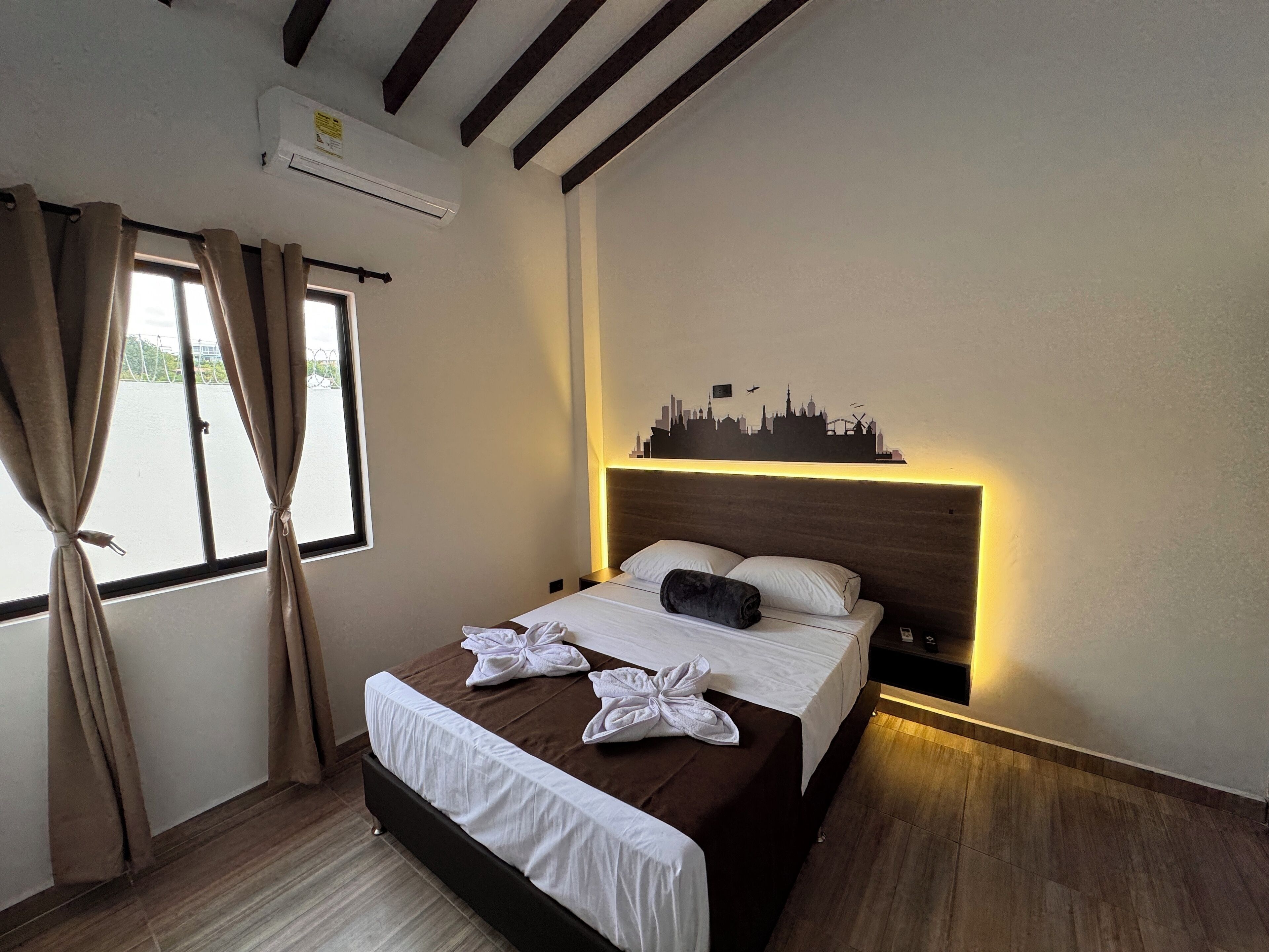 A hotel room with a bed, a cityscape wall art, an air conditioning unit, and a window with curtains.