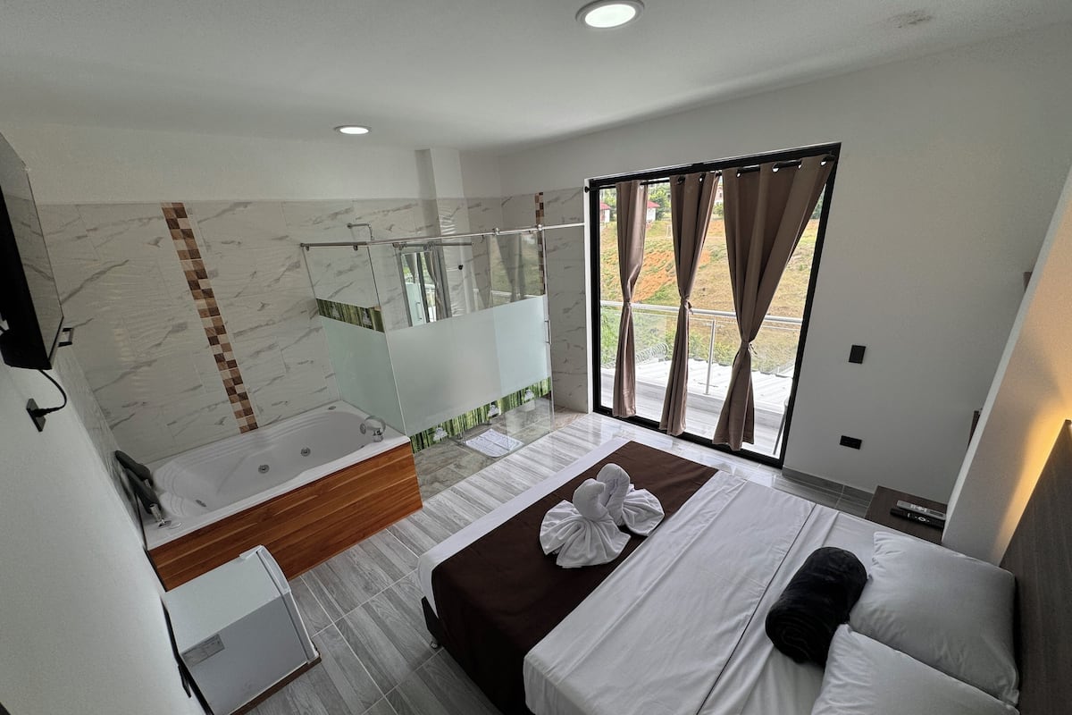 A modern bedroom with a large bed, a separate bathroom with a bathtub, and a view of the outdoors through a large window.
