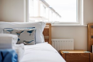 A neatly made bed with white bedding and a pillow featuring blue bird patterns, a wooden nightstand with a book, and a window with a view of a building. - Isle View - Large Corner Balcony With Harbour Views (Dorset)