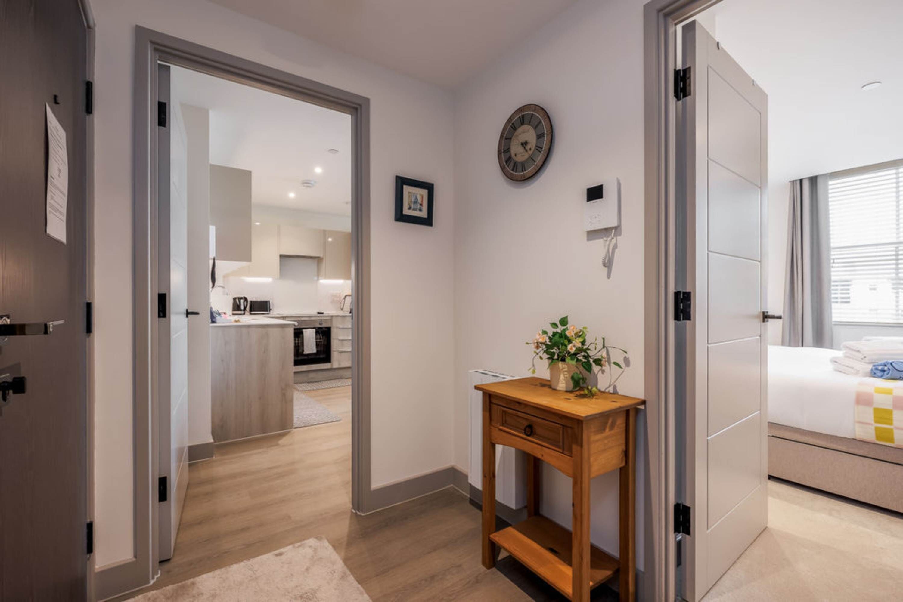 A modern hotel room with a wooden desk, a bedside table with a plant, a bed with a quilt, a clock on the wall, and a view of the kitchen and living area through an open door.