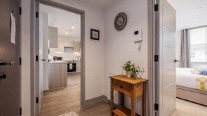 Une chambre d’hôtel moderne avec un bureau en bois, une table de chevet agrémentée d’une plante, un lit recouvert d’une courtepointe, une horloge au mur et une vue sur la cuisine et le salon, grâce à une porte ouverte.
