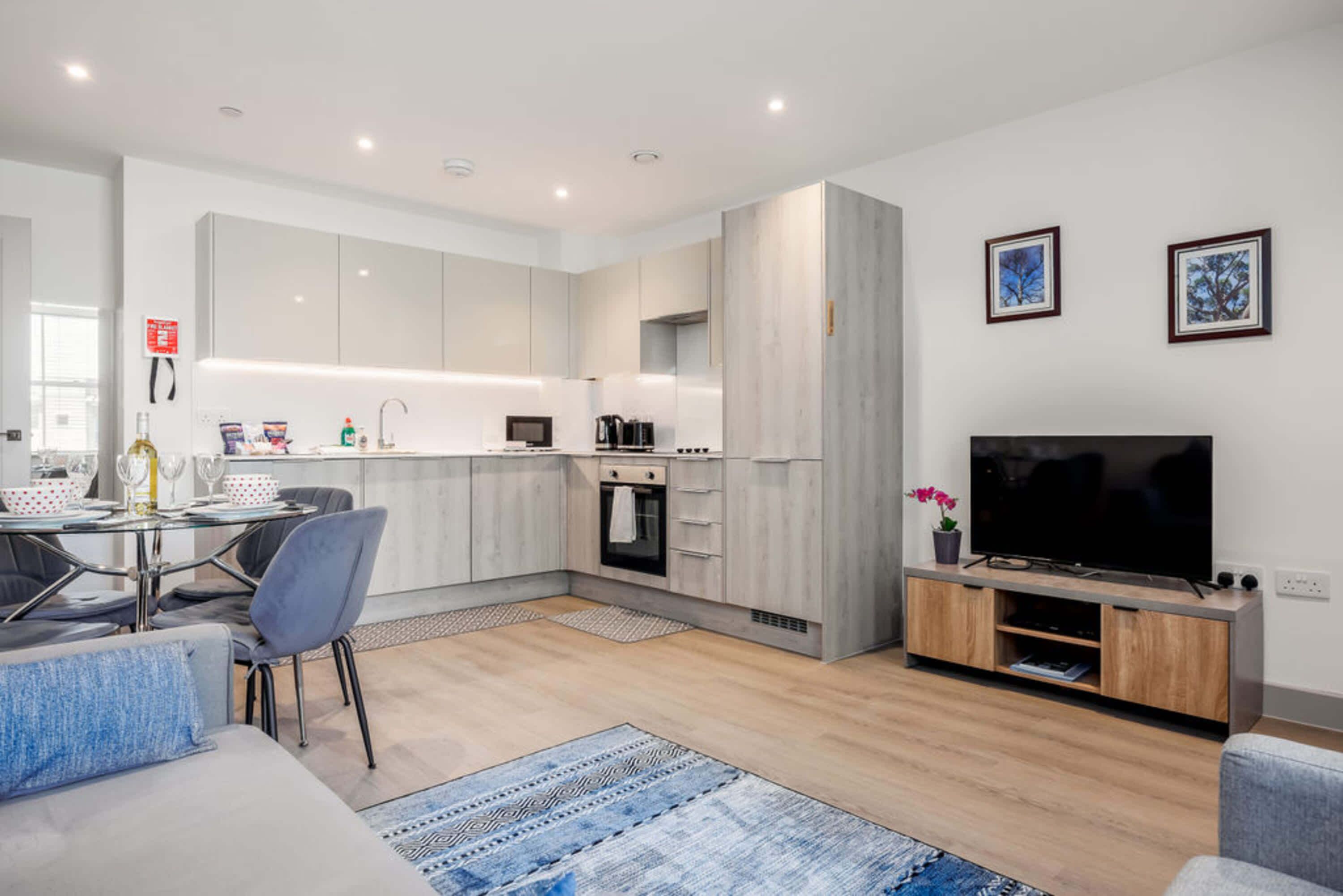 Una cocina moderna con armarios blancos, un comedor con mesa y sillas, un televisor de pantalla plana sobre un soporte de madera, y una sala de estar con sofá y alfombra azul.