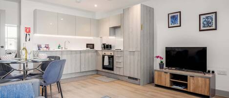 Una cocina moderna con armarios blancos, un comedor con mesa y sillas, un televisor de pantalla plana sobre un soporte de madera, y una sala de estar con sofá y alfombra azul.