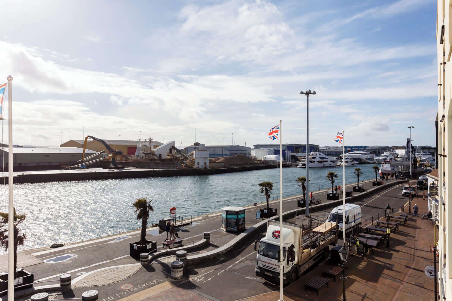 Un port de plaisance avec des bateaux, une route dotée d’une piste cyclable et un bâtiment sur lequel on peut voir une grue.