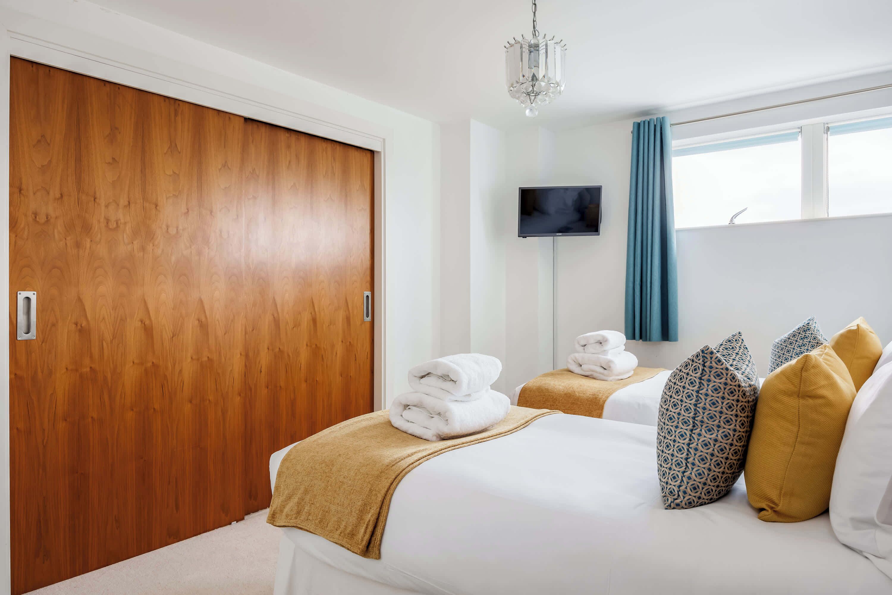 A hotel room with a wooden wardrobe, a television mounted on the wall, and two beds with towels and patterned pillows.