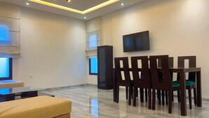 A modern living room with a glass coffee table, a sofa, a dining table with chairs, a TV mounted on the wall, and a tall cabinet. - Superb Apartment S+2 Sahloul 4 Sousse (Sousse)