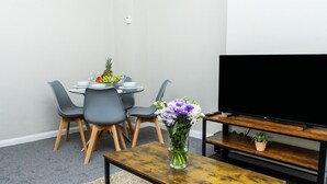 A dining area with a round table, grey chairs, a vase with flowers, and a TV on a wooden stand.