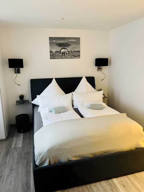 Un dormitorio moderno con cabecera negra, ropa de cama blanca, dos luces fijadas a la pared, una planta pequeña y un cuadro enmarcado de un paisaje.