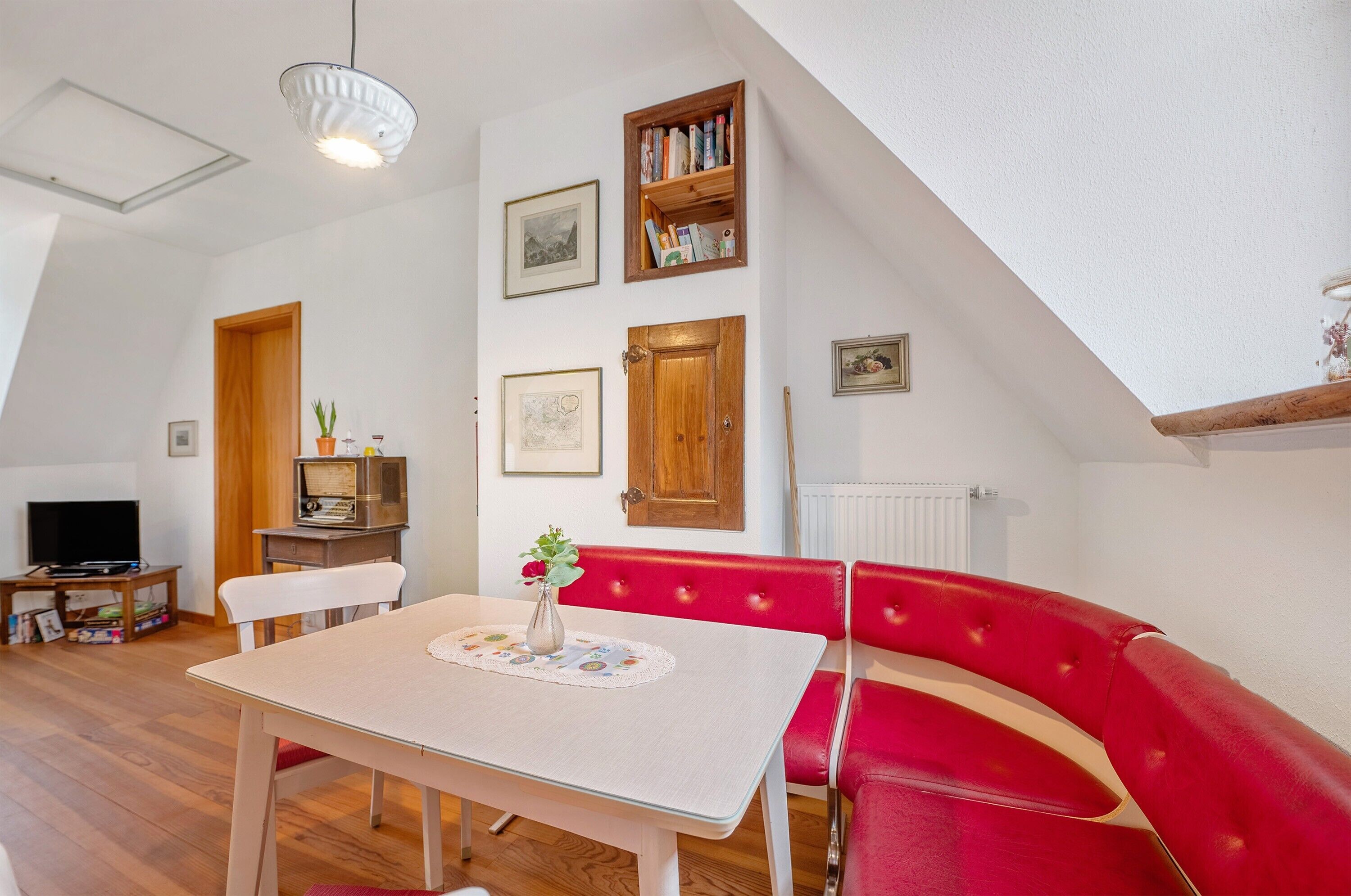 Ein Zimmer mit Esstisch, roter Sitzbank, weißen Wänden, Holztür, Bücherregal, gerahmten Bildern und einer Deckenleuchte.