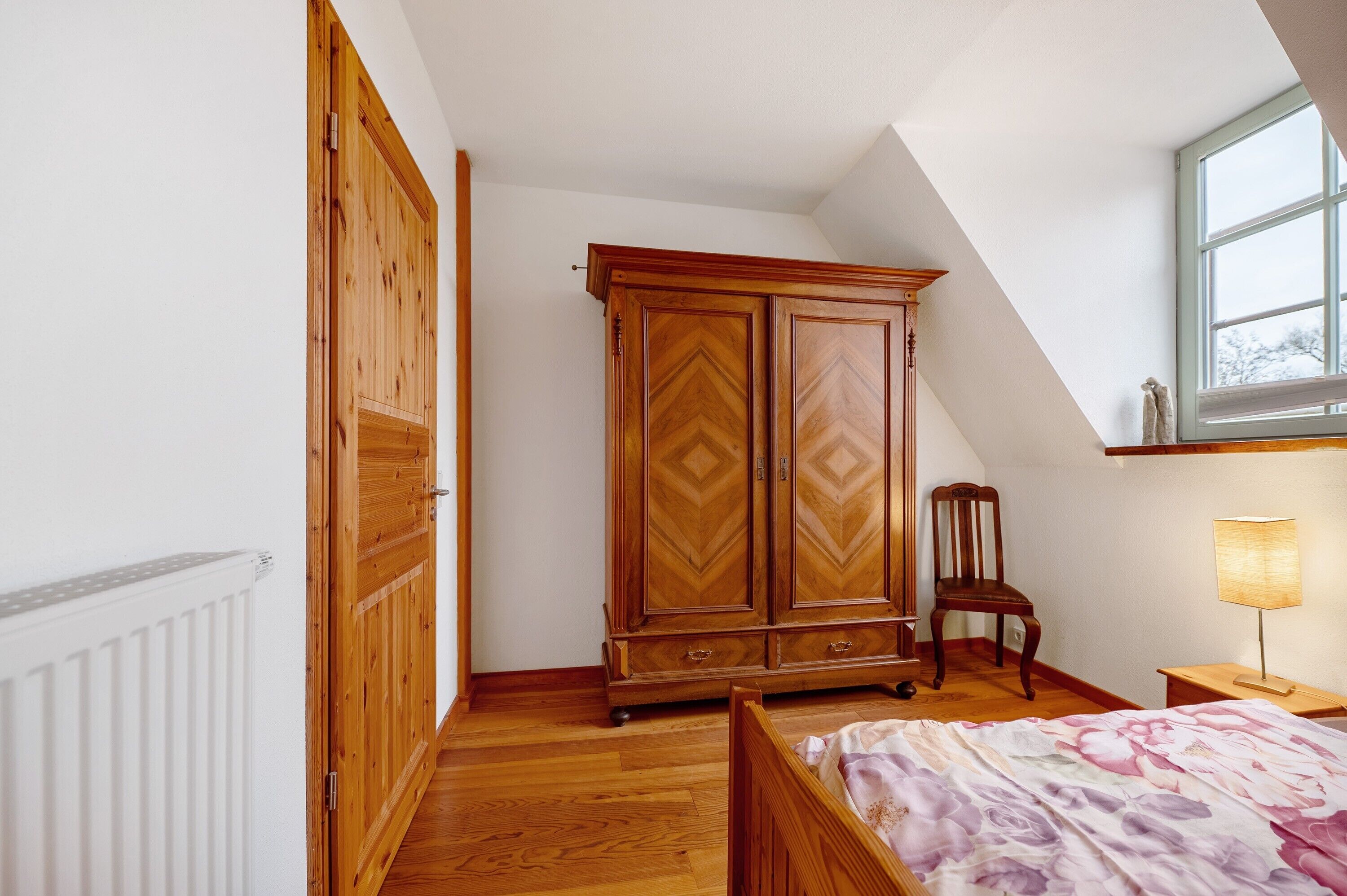 Une chambre avec une armoire en bois, un lit avec une literie à motifs floraux, une chaise, une table de chevet et une fenêtre donnant sur un beau paysage.