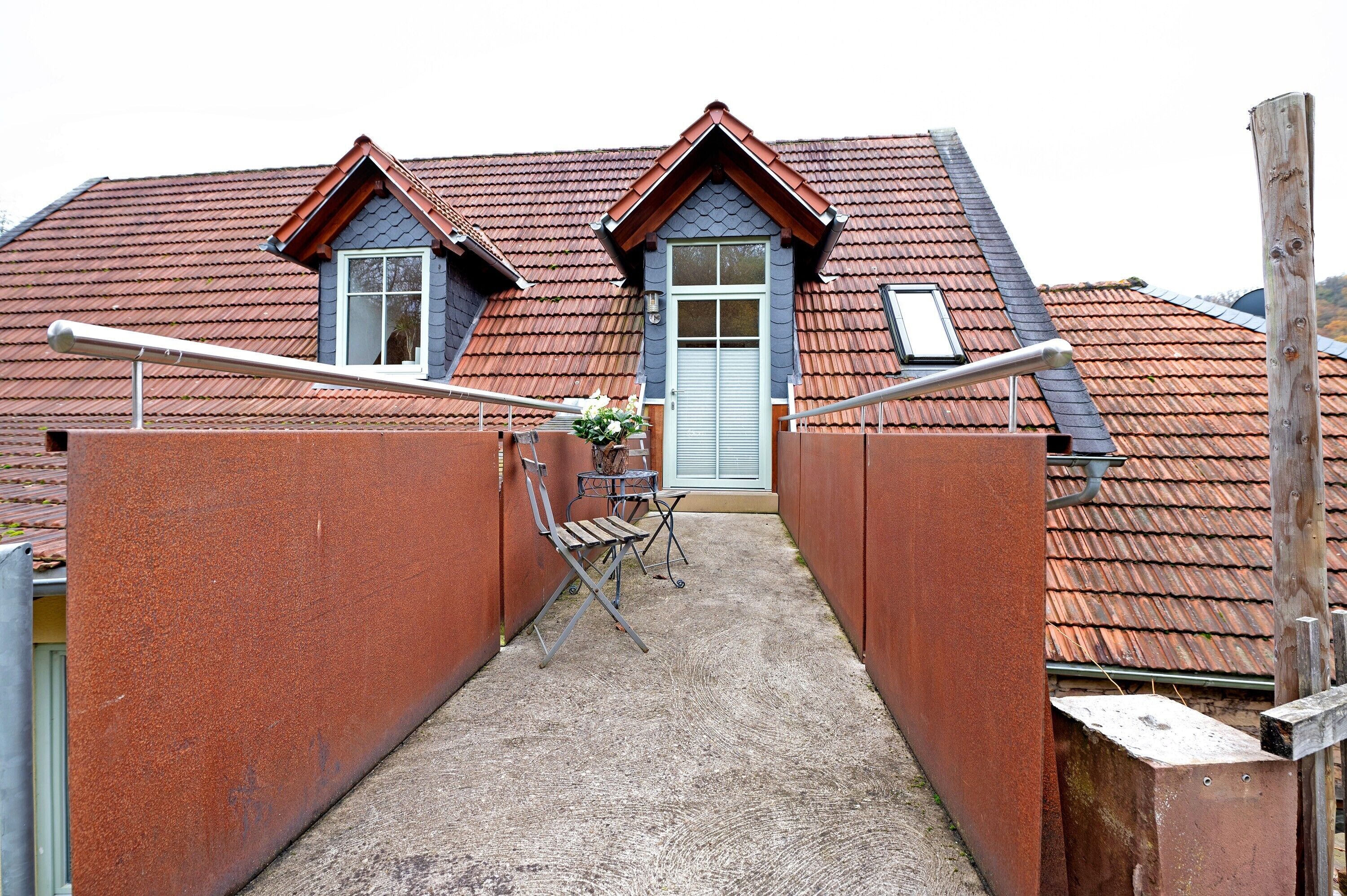 A narrow alleyway leading to a house with a red-tiled roof, a grey door, and a small table with chairs.