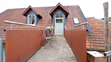 A narrow alleyway leading to a house with a red-tiled roof, a grey door, and a small table with chairs.