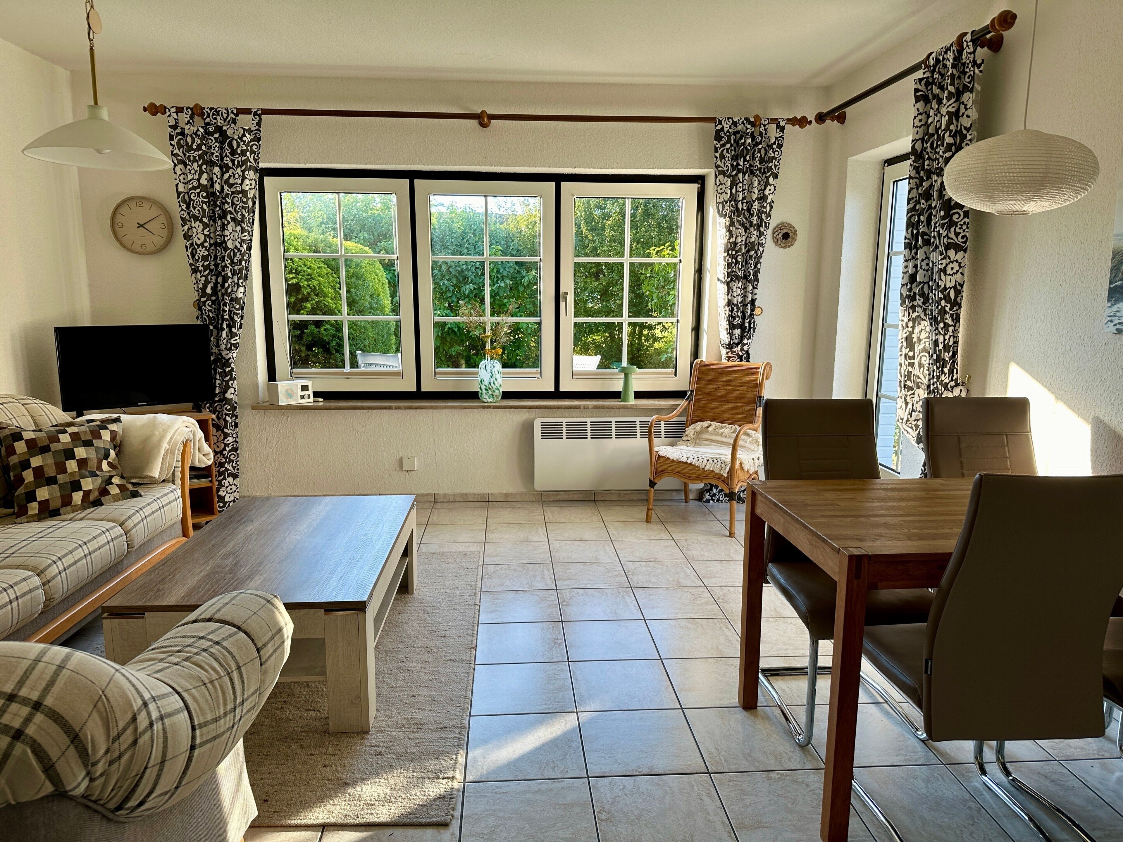 A living room with a sofa, a wooden table, a chair, a television, and large windows with curtains.