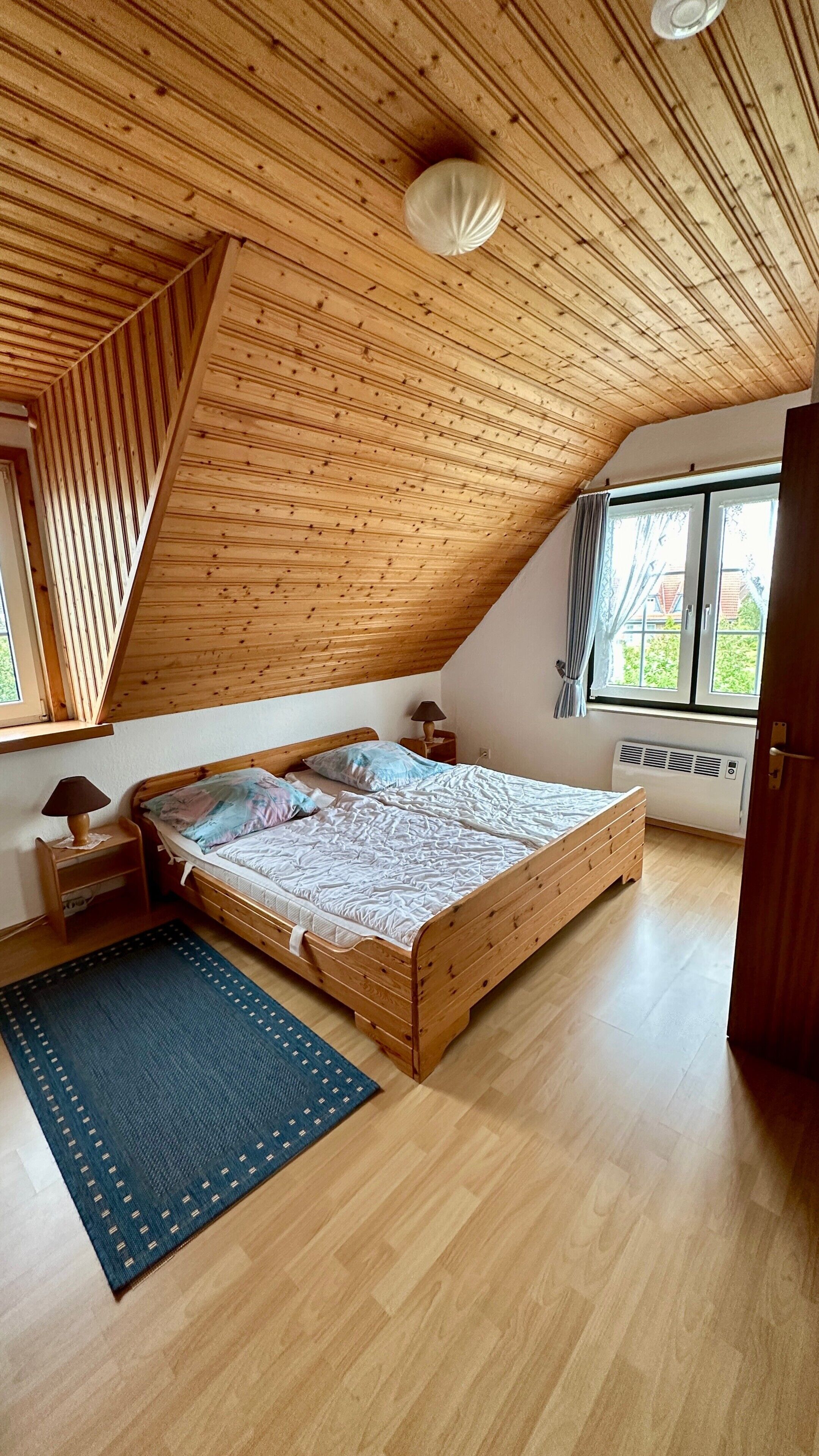 Une chambre avec un plafond en bois incliné, un lit, une fenêtre et un radiateur.