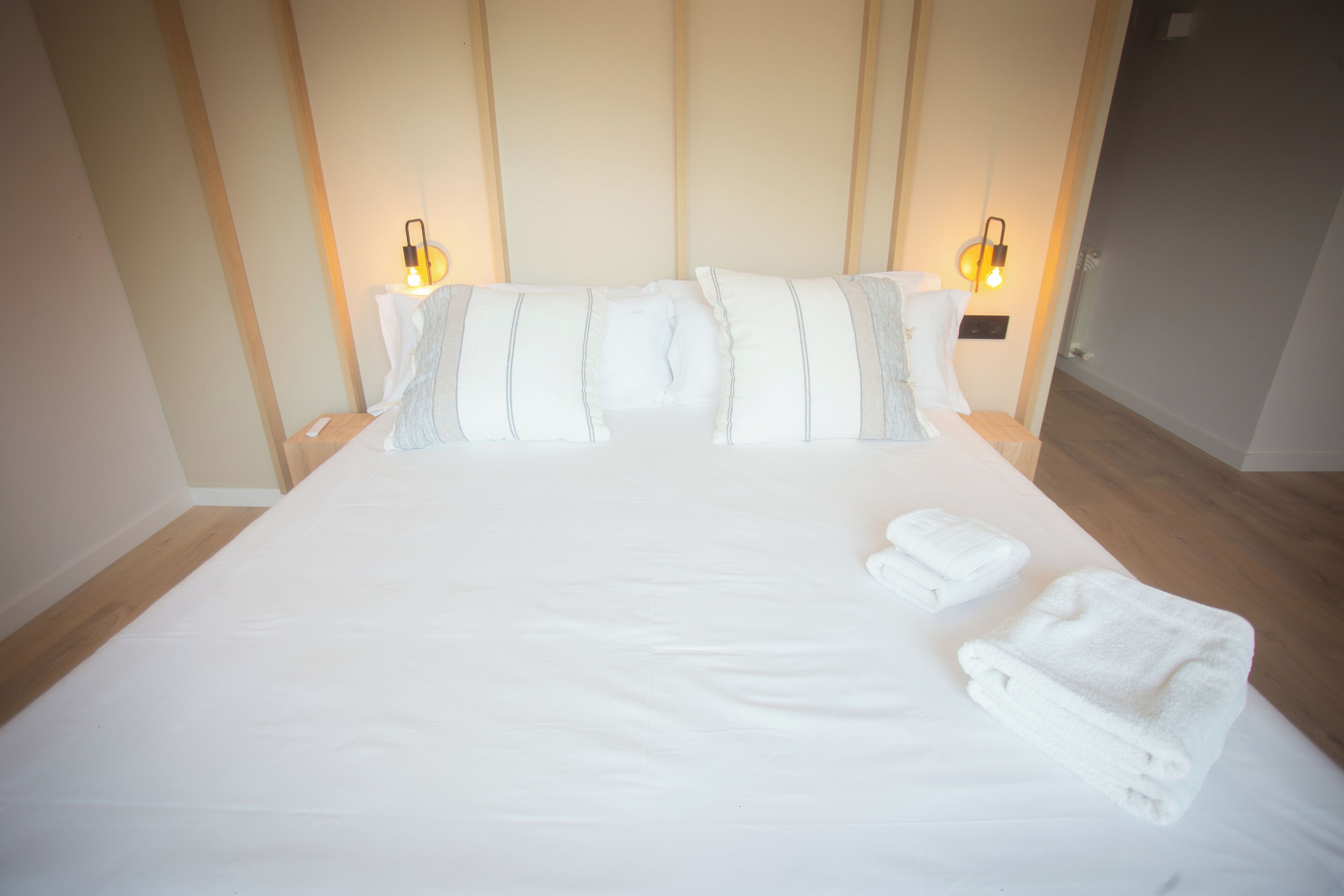 Una cama bien hecha con sábanas blancas y dos almohadas, dos lámparas de noche y dos toallas dobladas sobre la cama.