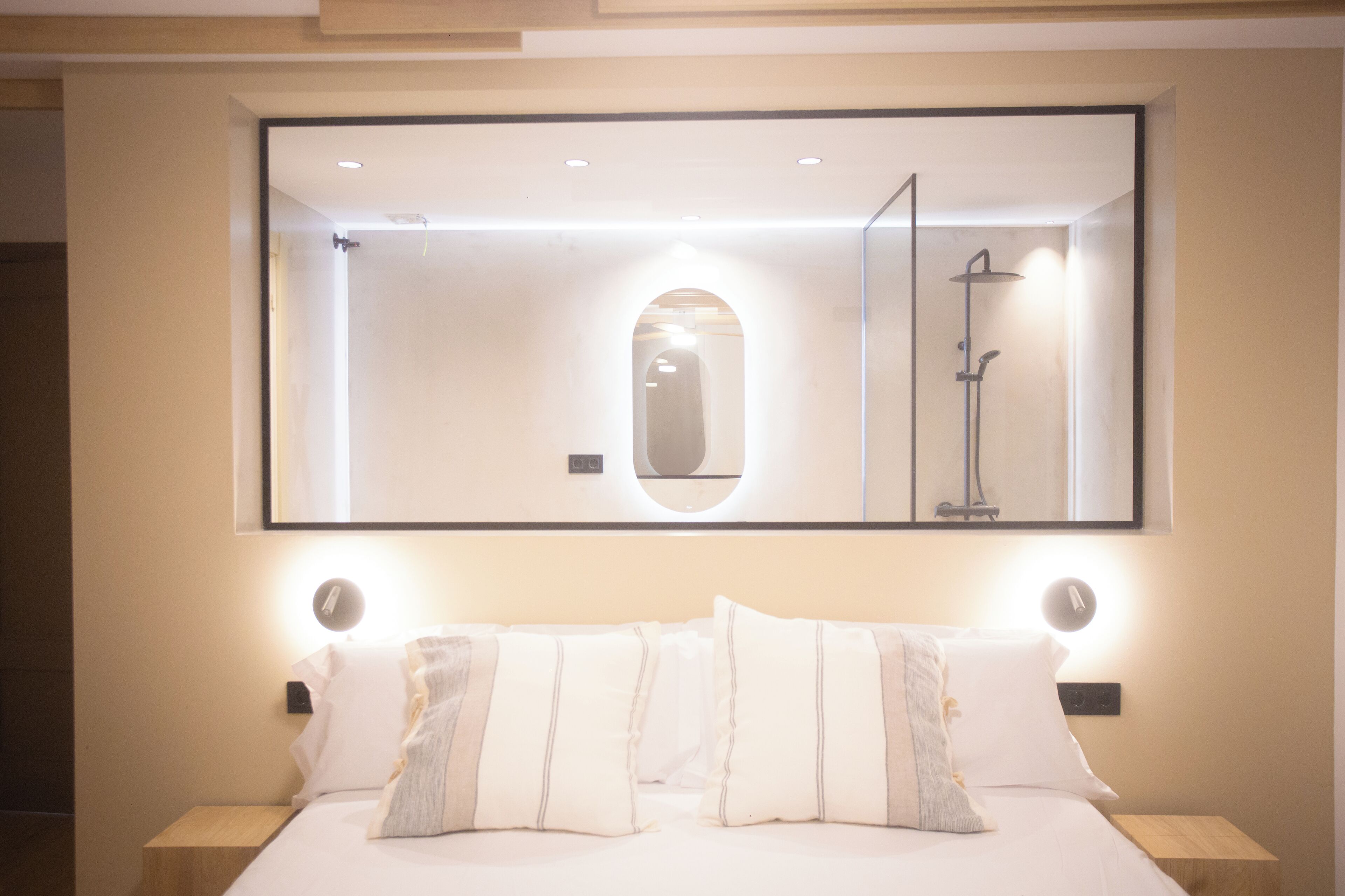 A hotel room with a large mirror above the bed, a nightstand with a lamp, and a shower visible in the mirror reflection.