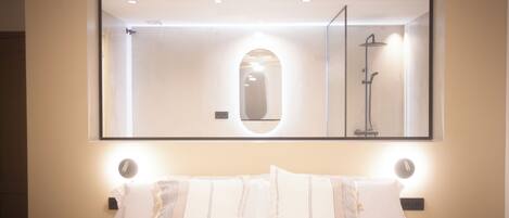 A hotel room with a large mirror above the bed, a nightstand with a lamp, and a shower visible in the mirror reflection.