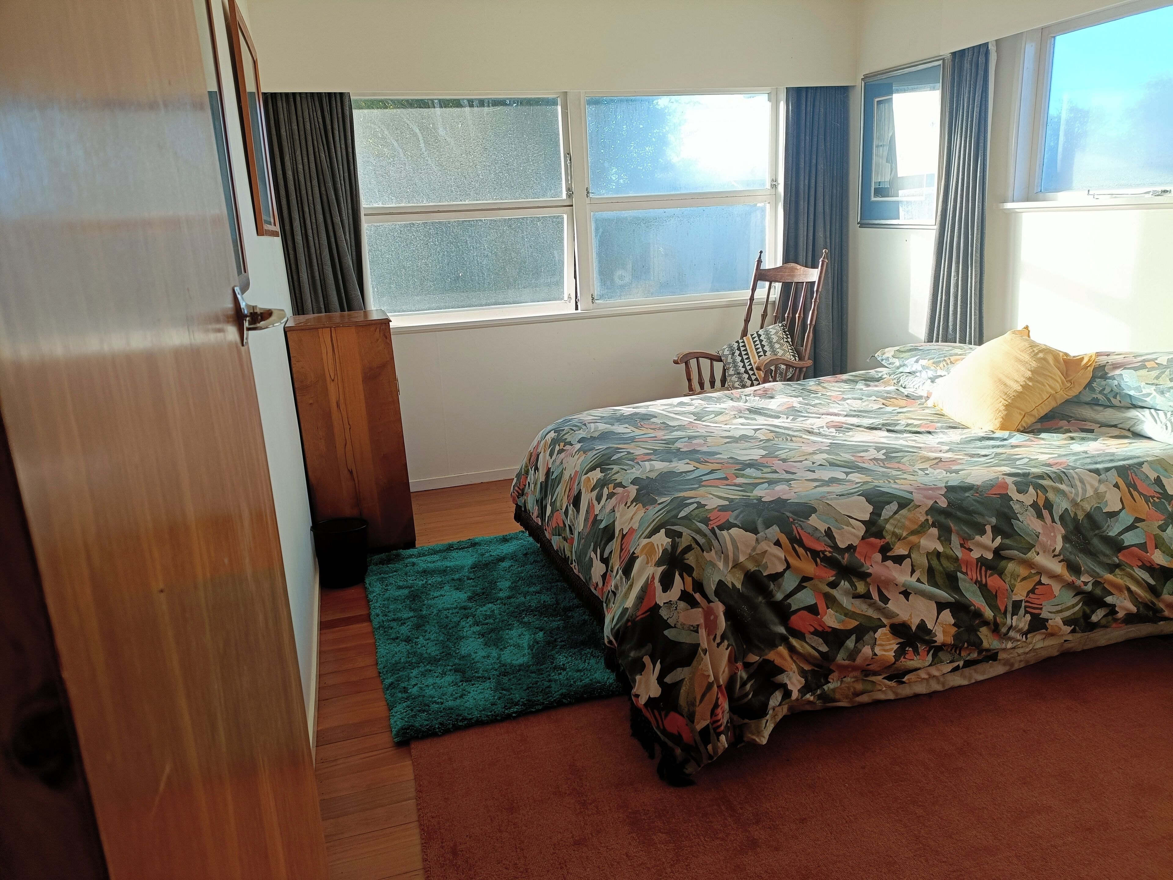 A bedroom with a bed, a wooden chair, a green rug, and a window with curtains.