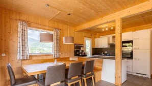 A wooden cabin interior with a dining area, kitchen, and large windows offering a mountain view. - Modern chalet with infrared sauna (Sankt Georgen am Kresichberg)