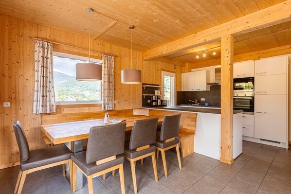 Interior de una cabaña de madera con comedor, cocina y ventanas grandes que ofrecen vistas a la montaña.
