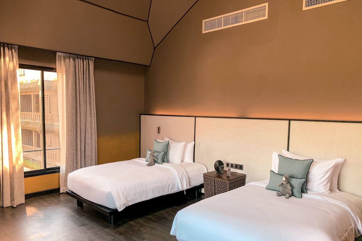 Ein Hotelzimmer mit zwei Betten, einem Fenster mit Vorhängen, einem Nachttisch und einem Wandlüfter.