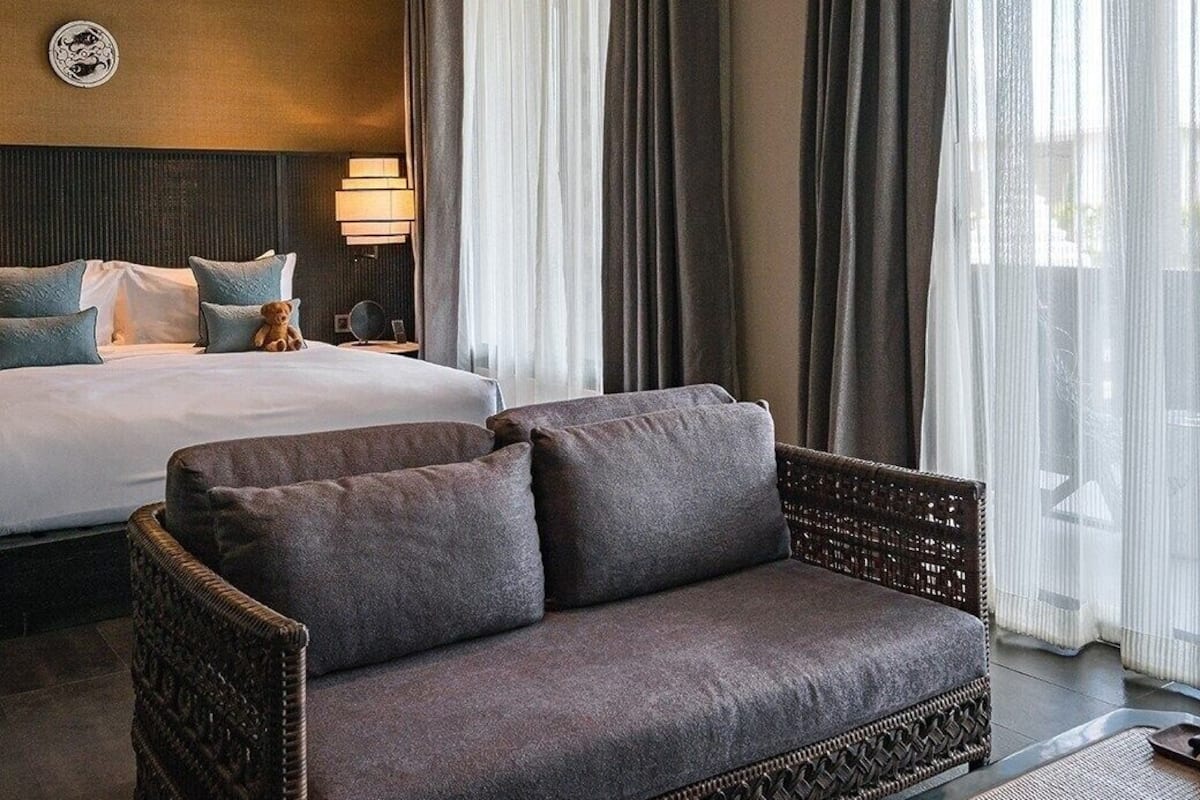 Ein Hotelzimmer mit Bett, einer Couch, einer Lampe und Blick ins Freie durch transparente Vorhänge.