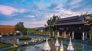 Un étang paisible, parsemé de fleurs de lotus et bordé de pagodes illuminées, est entouré d’un bâtiment moderne à la façade en bois et d’une structure traditionnelle surmontée d’un toit en tuiles.