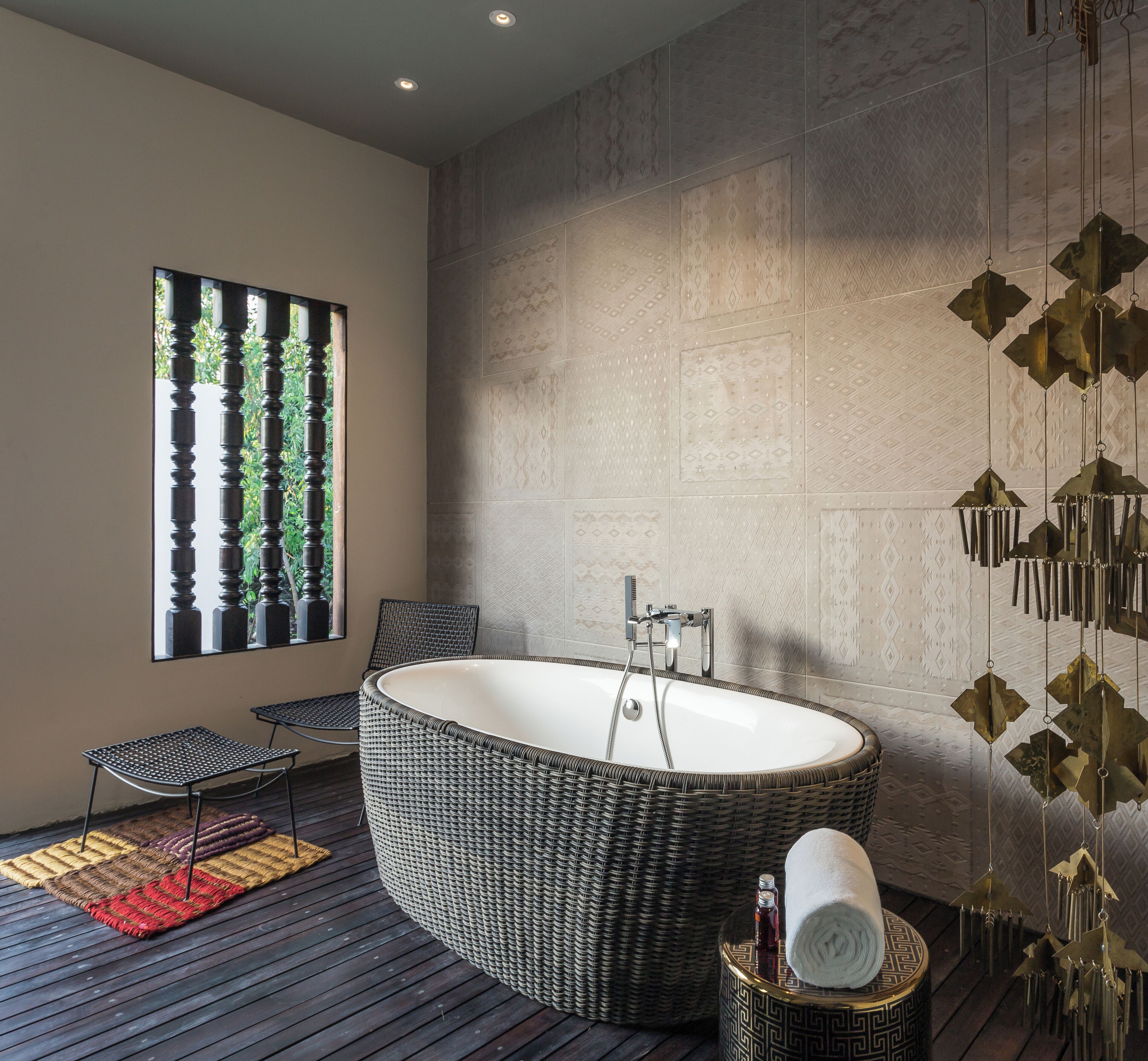 A modern bathroom with a freestanding bathtub, a window with vertical blinds, a wicker chair, and a decorative wall hanging.