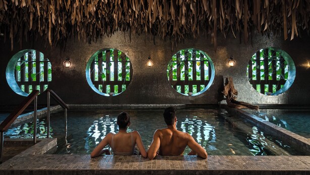 Un couple est assis au bord d’une piscine, à côté d’un bâtiment dont le toit est fait de chaume et qui possède des fenêtres rondes.