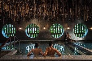 Una pareja sentada junto a una piscina con techo de paja y ventanas circulares.