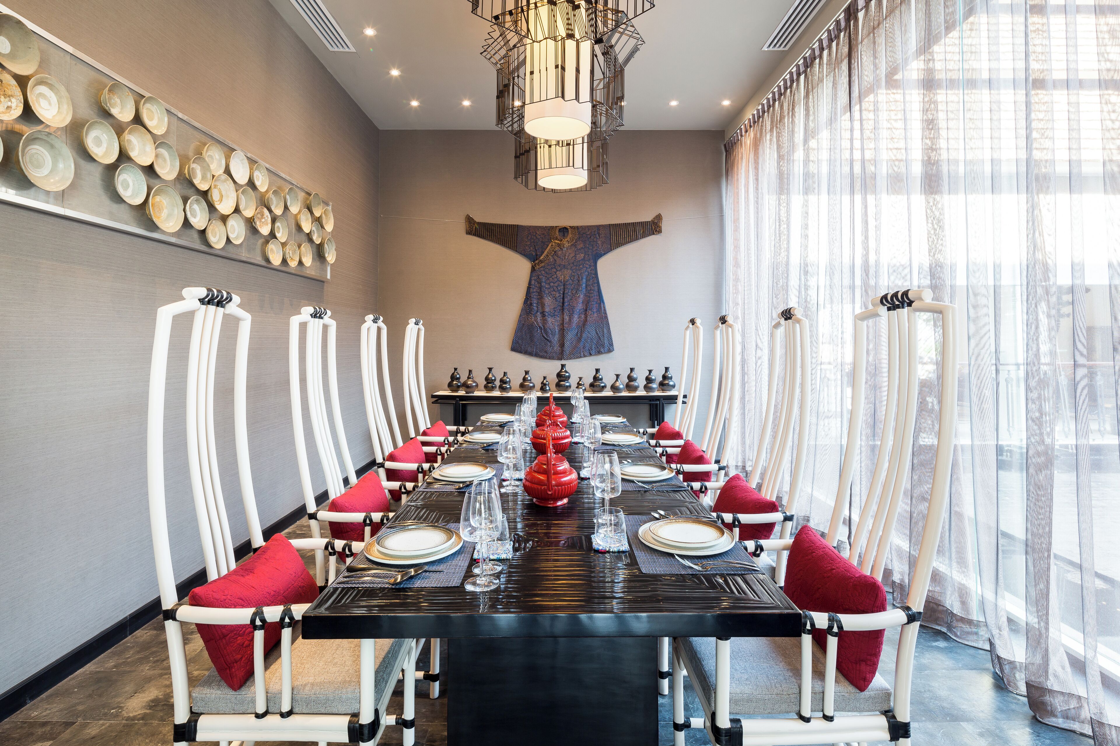 A dining room with a long table set for a meal, white chairs with red cushions, a chandelier, and a wall with a tapestry.