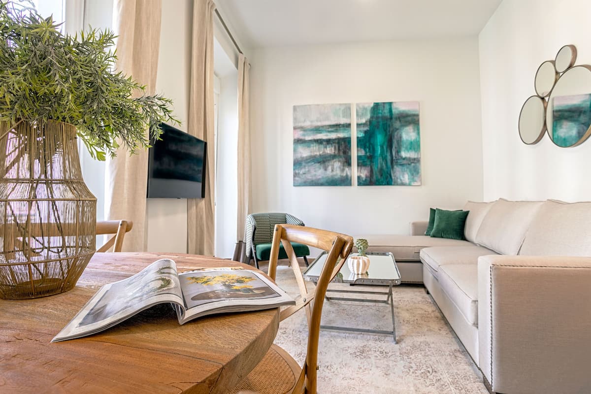 Un salon avec une table en bois, un fauteuil vert, un canapé beige et des œuvres d’art abstraites accrochées au mur.