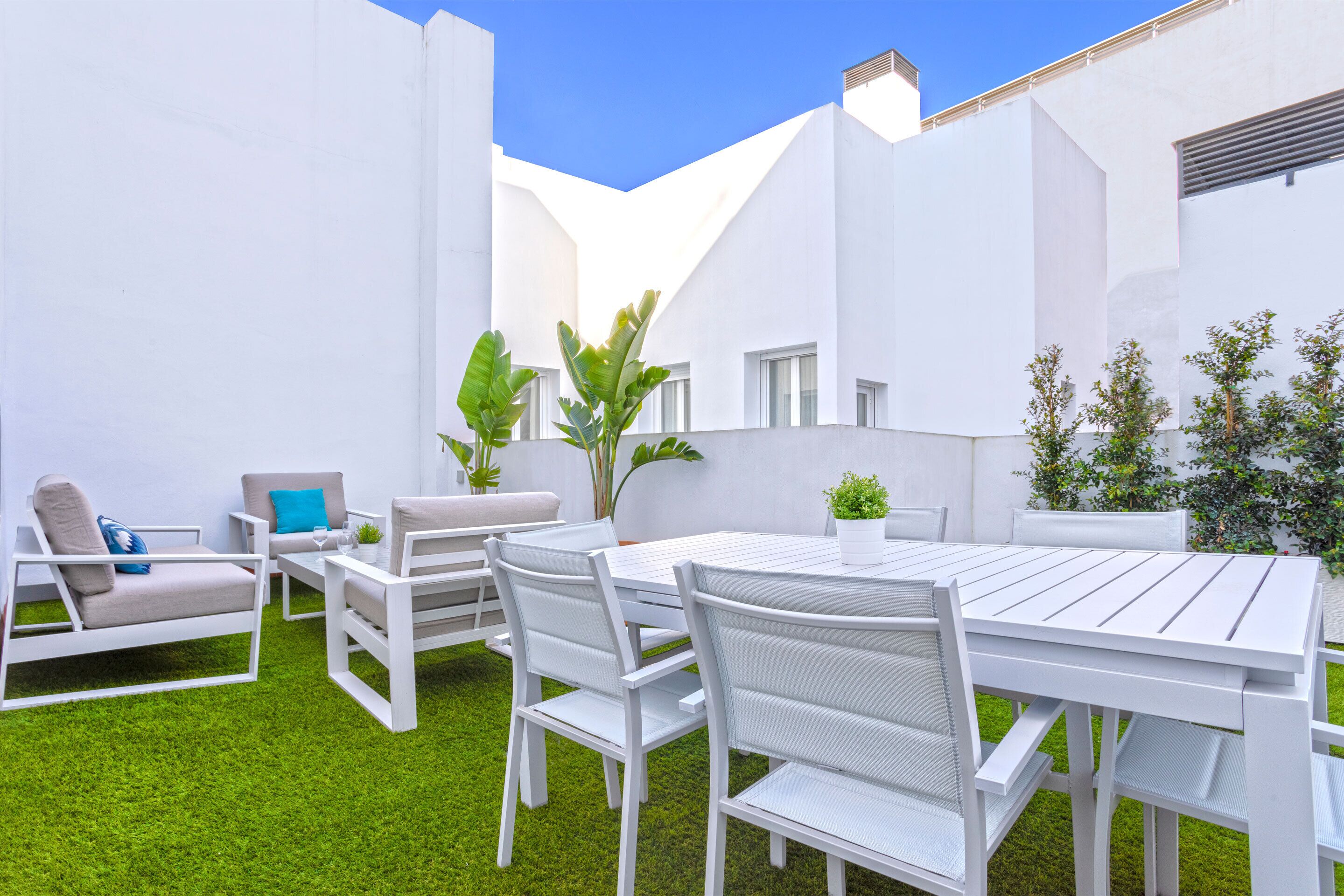 Een wit terras buiten met een tafel en stoelen, omringd door witte muren en groen gras.