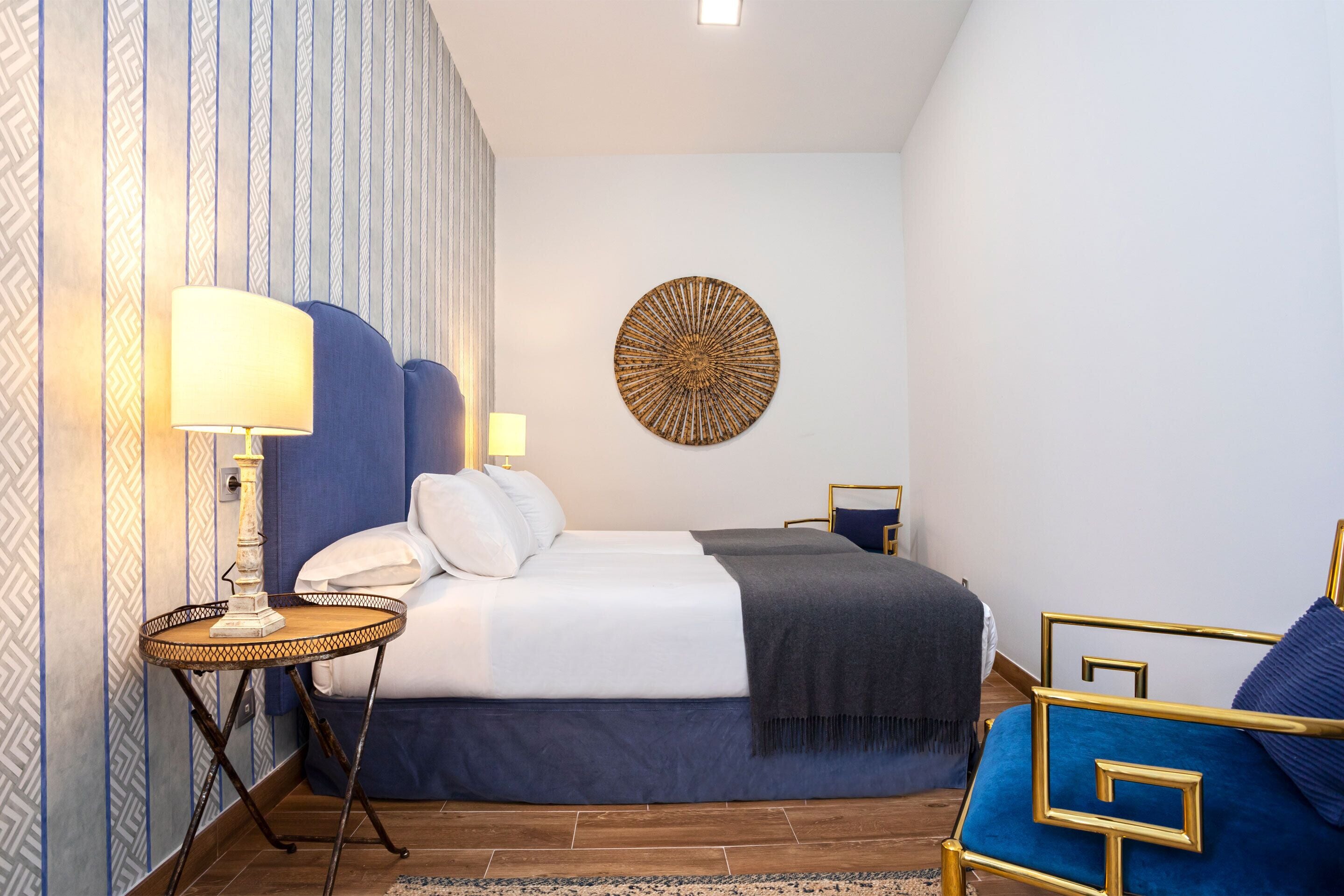 Habitación de hotel con una cama grande, una mesita de noche con lámpara, un adorno en la pared y un sillón azul.