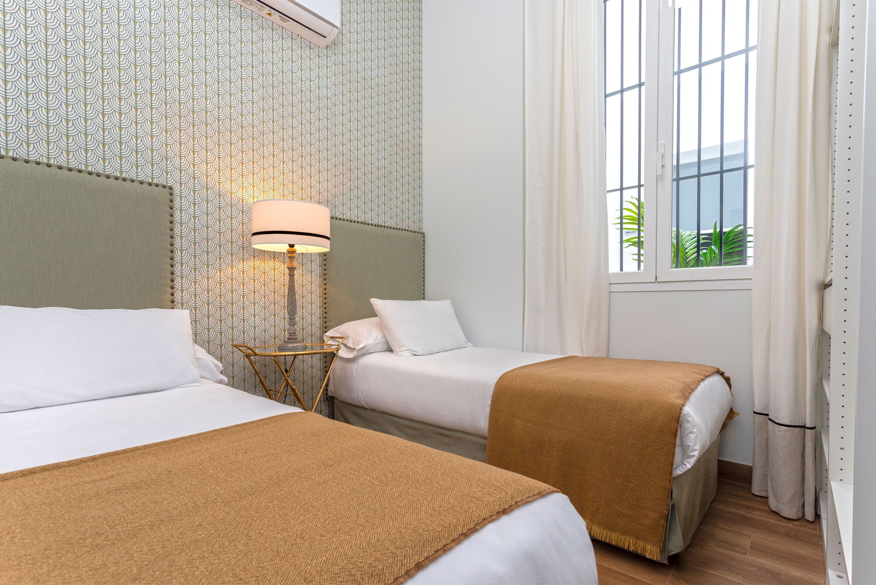 A hotel room with two beds, a lamp, a window with curtains, and patterned wallpaper.