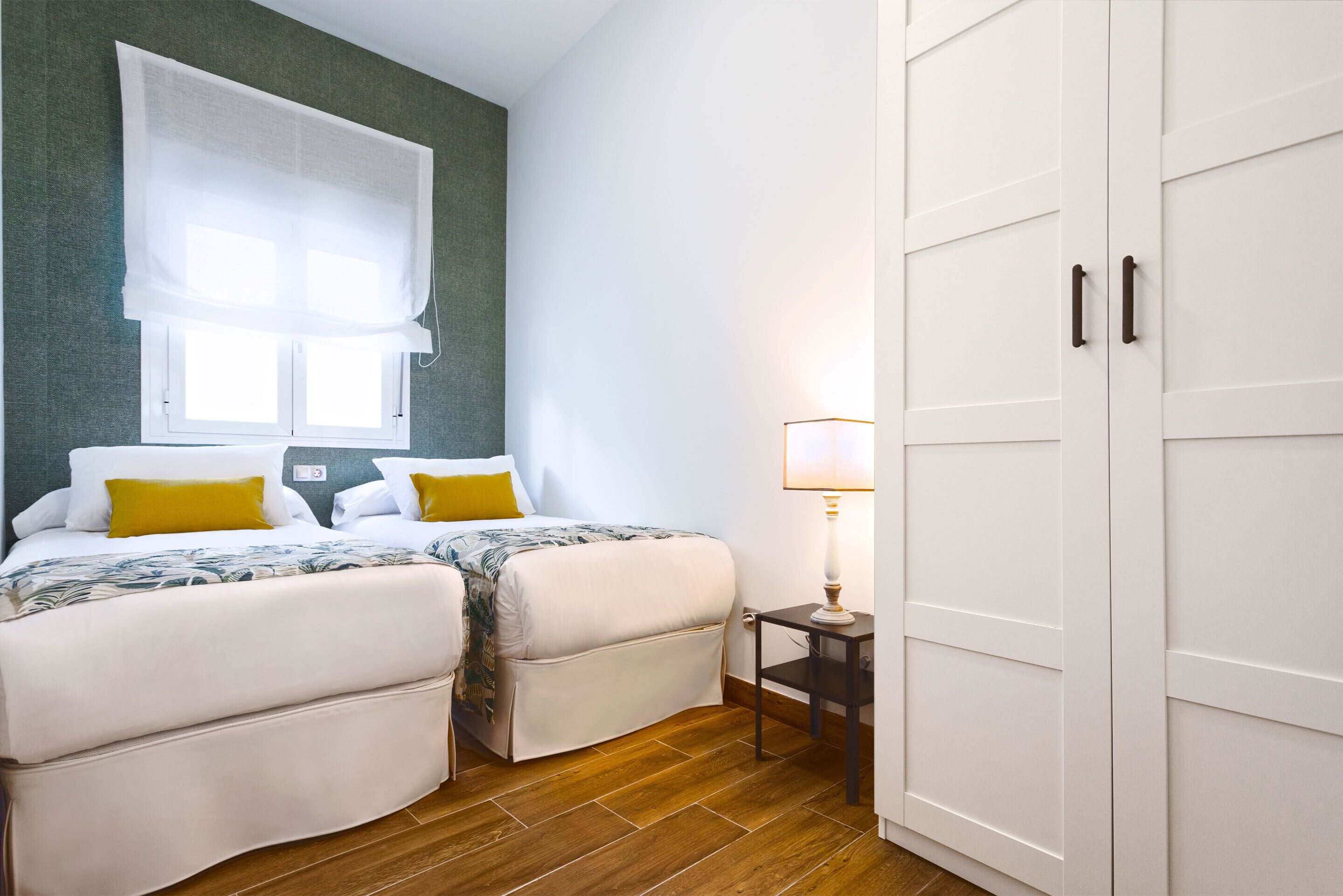 Une chambre avec deux lits, une fenêtre avec des rideaux blancs, une table de chevet avec une lampe et une armoire.