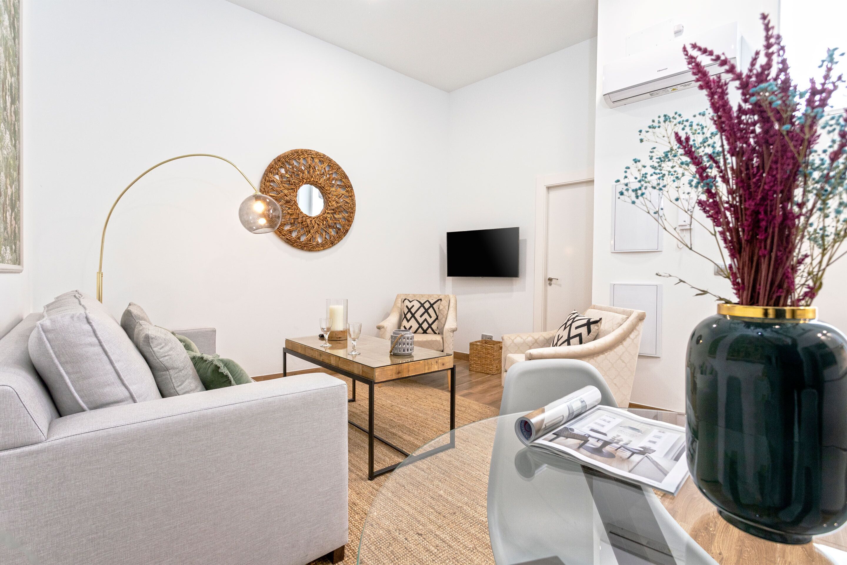 A modern living room with a sofa, coffee table, and chairs. A flat-screen TV is mounted on the wall.
