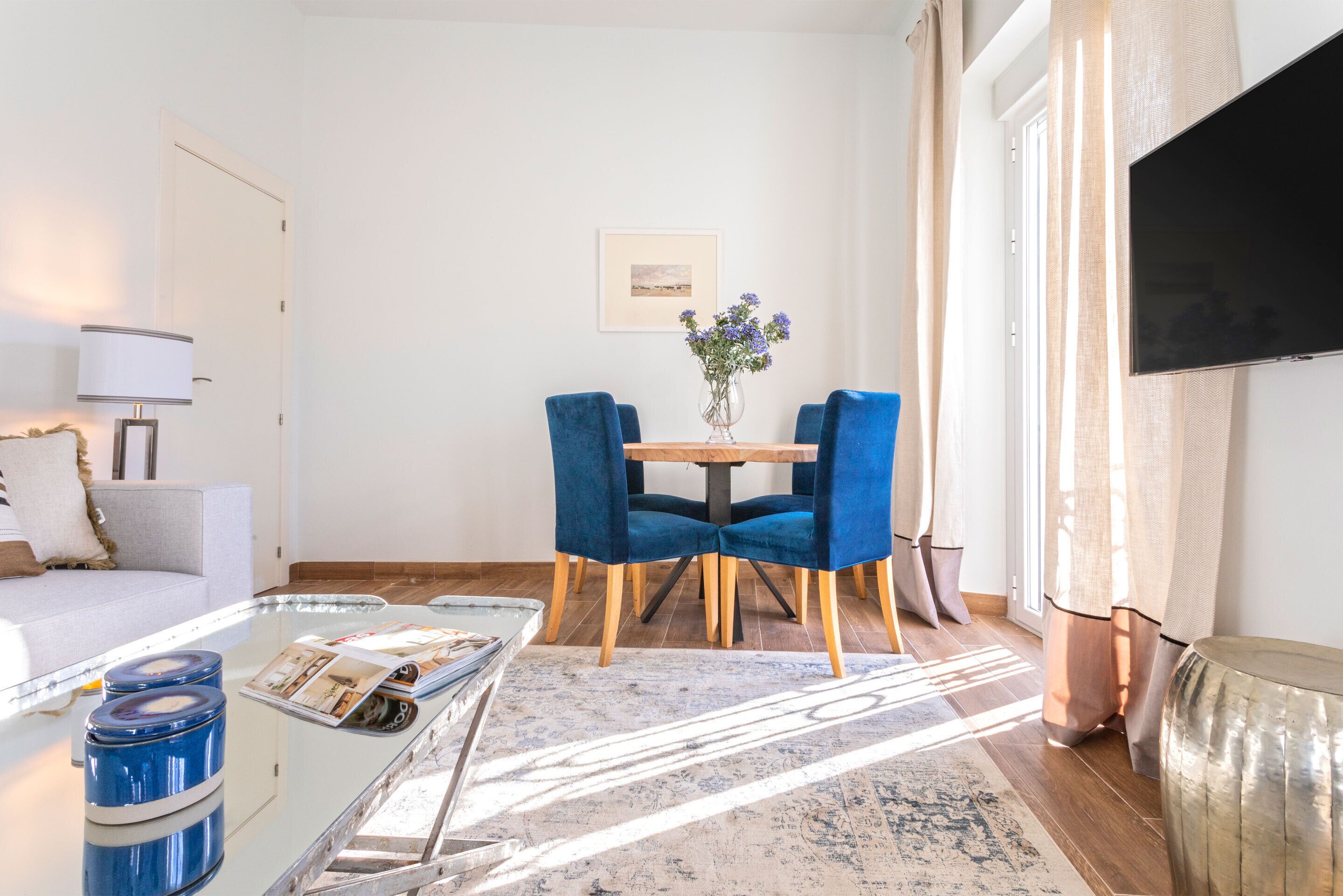 Una sala de estar moderna con un sofá, una mesa de centro, una mesa de comedor con sillas azules, un televisor montado en la pared y una ventana con cortinas.