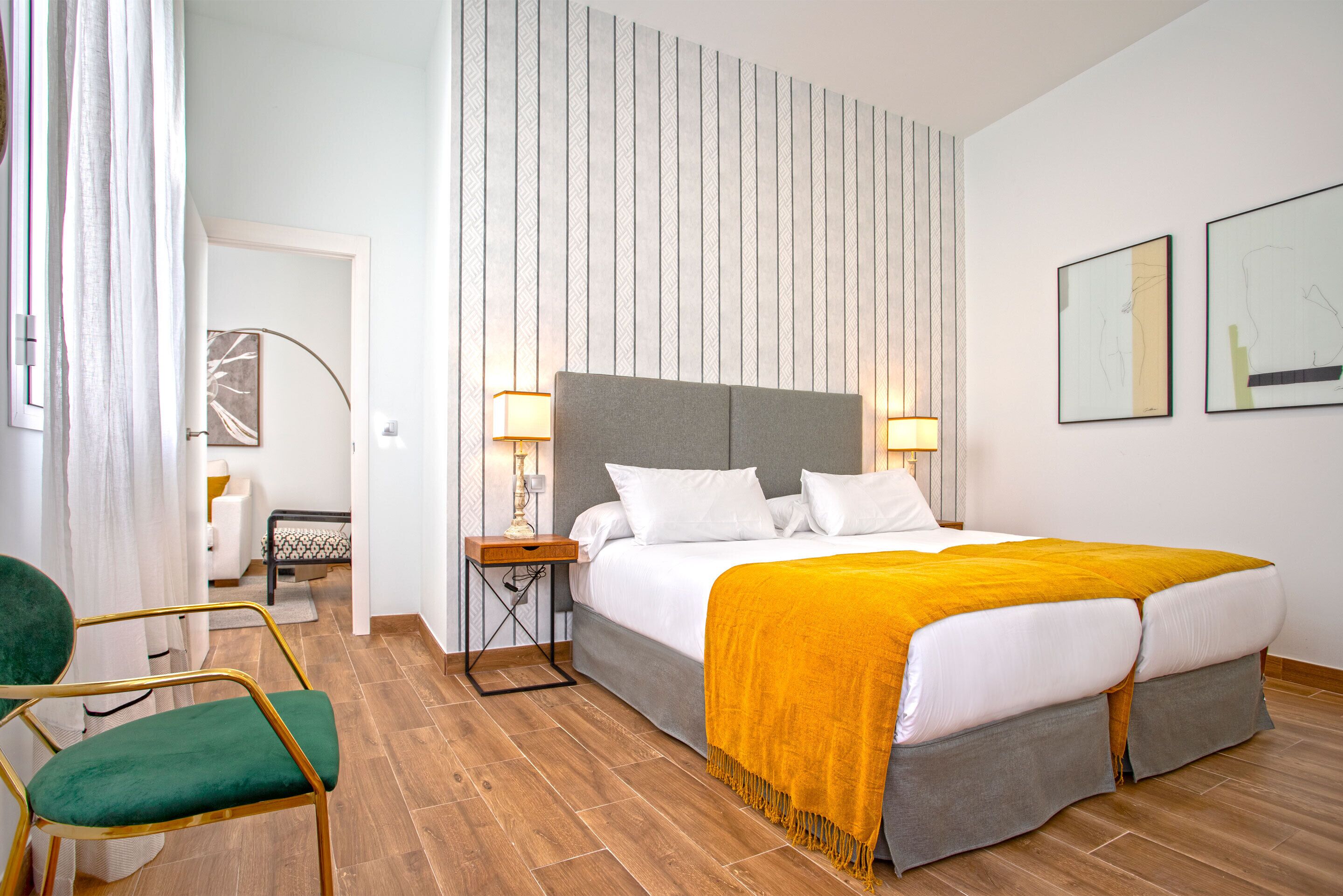 A hotel room with a double bed, two bedside lamps, a green chair, a small table, and a decorative wall panel.
