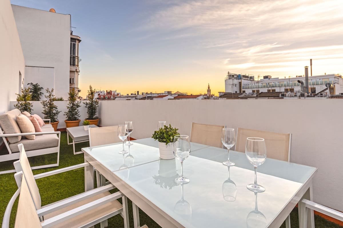 Une terrasse sur le toit avec une table blanche dressée pour le dîner, entourée de chaises blanches, et une petite plante en pot.