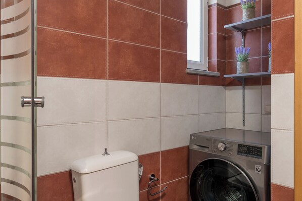 A compact laundry room with a washing machine, a toilet, and a shelf with potted plants. - Olive Tree Villa (Chania)