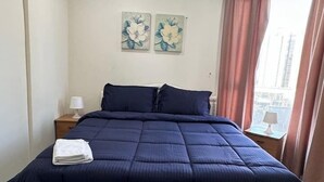 Un letto rifatto con un copriletto blu scuro e cuscini, una lampada bianca su un comodino e due dipinti floreali al muro.