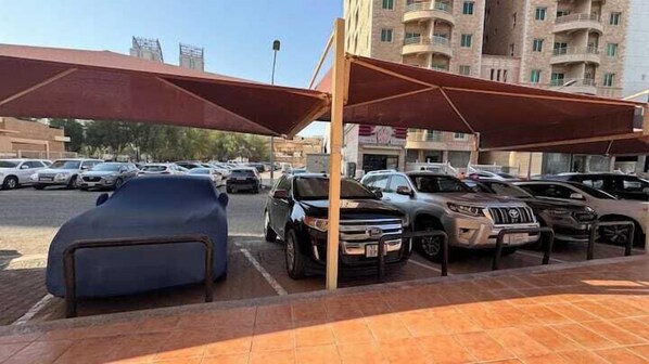 A parking area with multiple cars, a building with balconies in the background, and a clear sky. - Basser 6 (Salmiya)